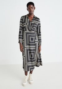 Abito lungo avvolgente in bianco e nero, con motivi geometrici a quadrato. Realizzato in tessuto leggero, con colletto e dettagli con bottoni.