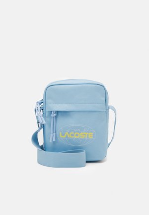 Sac bandoulière bleu clair avec poche zippée à l'avant et logo Lacoste jaune sur un motif carte du monde à l'avant.