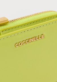 Vihreä nahkalompakko, jossa on teksturoitu pinta, sileä vetoketju ja kullanväriset logoyksityiskohdat. Vetoketjussa on samansävyinen korostus.