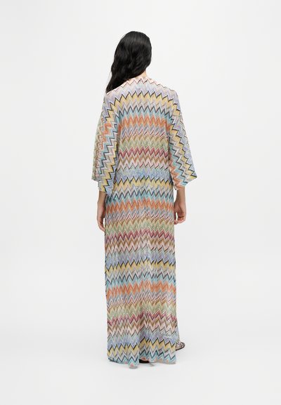 Femme aux longs cheveux noirs portant une longue robe ample à motif zigzag multicolore, debout de dos sur un fond blanc uni.