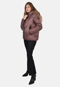 Giacca imbottita marrone trapuntata con collo alto, tasche con zip e toppa con logo sulla manica. Indossata sopra jeans neri e stivali con punta affilata.