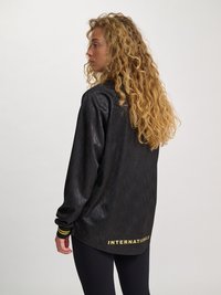 Kvinne med langt krøllete blondt hår har på seg en svart langermet jakke med "INTERNATIONALS" trykket i gult på bakre kant.