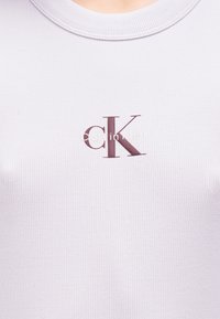 Ανοιχτό μωβ ριμπ αμάνικο τοπ με διακριτικό λογότυπο "CK" σε μπορντό και λευκά γράμματα στην πε ριοχή του στήθους.