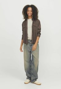 Camicia marrone a bottoni sopra una maglietta beige, abbinata a jeans chiari e larghi e sneakers beige. Capelli acconciati in ricci naturali.