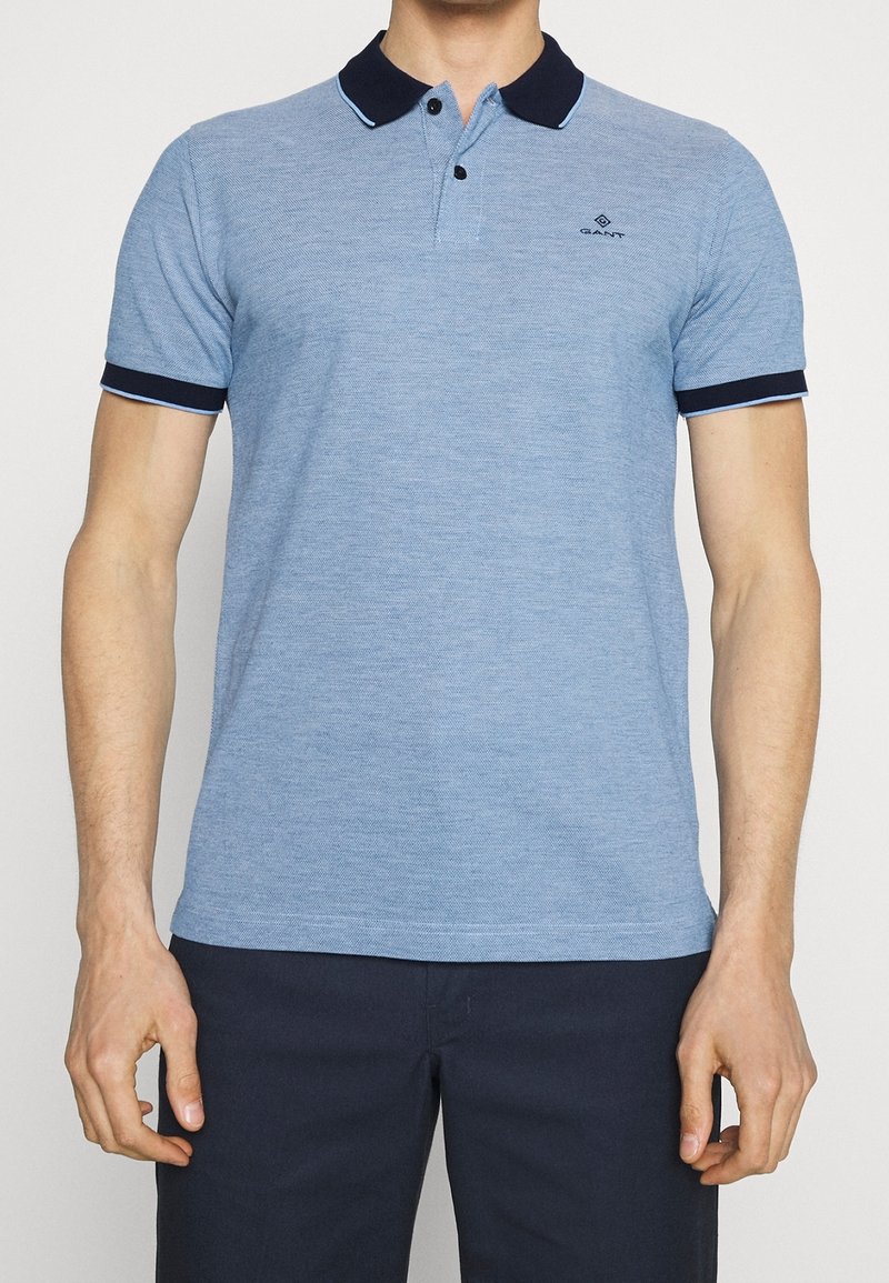 Homme portant un polo à manches courtes bleu clair avec un col et des bords de manches bleu foncé, associé à un pantalon bleu foncé, debout droit.