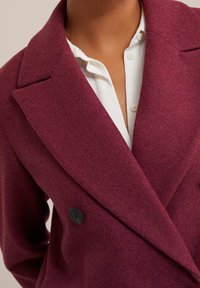 Cappotto doppiopetto in misto lana color bordeaux con risvolti a lancia, caratterizzato da un bottone nero e texture morbida, abbinato a una camicia bianca sotto.