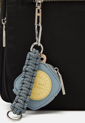 Personne portant un haut noir et un jean cargo bleu, portant un sac à dos noir avec une petite pochette grise attachée.