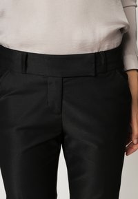 Svarta skräddarsydda byxor med en slät yta, rak framdel, sidofickor och ett robust midjeband. Bärs med en ljusfärgad topp.