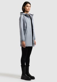 Frau trägt eine hellgraue Kapuzenjacke, schwarze Leggings und schwarze klobige Stiefel, steht in neutraler Haltung vor einem schlichten Hintergrund.