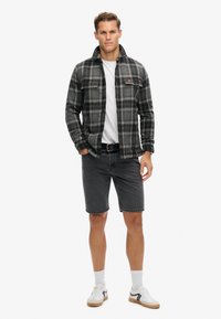 Veste en flanelle à carreaux noirs avec deux poches poitrine, t-shirt blanc à col rond, shorts en denim gris et baskets blanches avec accents noirs.