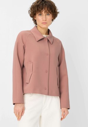 Woman with short curly hair wearing a muted pink buttoned jacket with two front pockets and white pants, standing against a plain background.