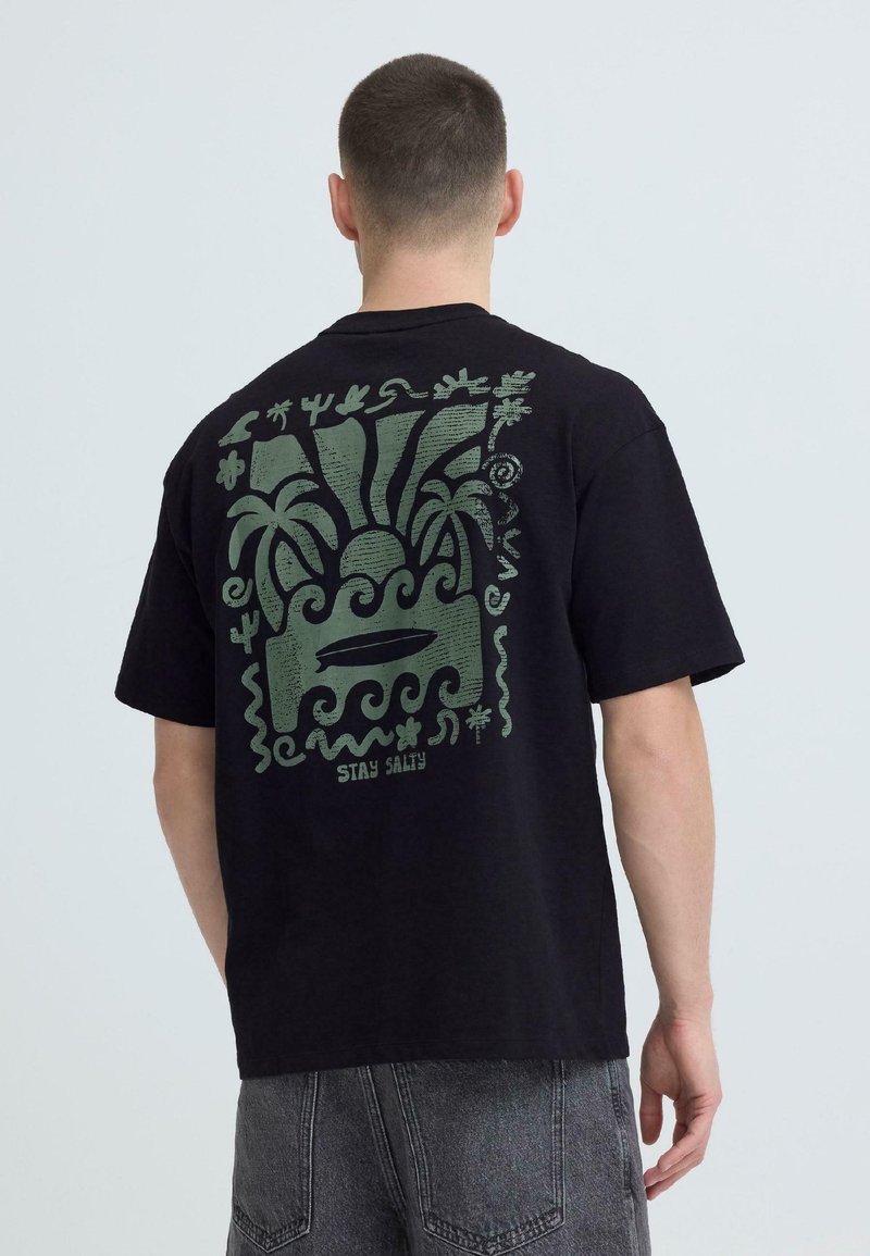 Άνδρας που φοράει μαύρο μπλουζάκι με πράσινο γραφικό ήλιο, κύματα, φοίνικες, κάκτους και σανίδα του σερφ, με το κείμενο «Stay Salty» στην πλάτη.
