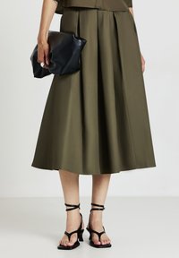 Gonna A-line khaki realizzata in tessuto morbido, caratterizzata da delicate pieghe. Abbinata a una borsa a tracolla nera in pelle e tacchi neri con cinturini.