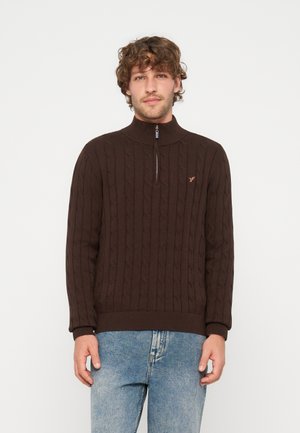 Jeune homme aux cheveux bouclés portant un pull marron en maille torsadée avec fermeture éclair mi-longue et un jean bleu, debout devant un fond blanc uni.