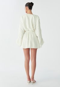 Een vrouw met een strakke bun draagt een witte, geplooide mini-jurk met lange mouwen terwijl ze tegen een lichte achtergrond staat.