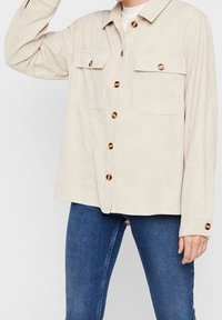 Beige kordflodskjorta med bruna knappar, bärs över en vit topp, tillsammans med blå denimjeans. Visar överkroppen med armen höjd.