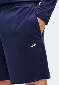 Homme portant un short de sport bleu marine avec une ceinture élastique et un logo Reebok blanc sur la cuisse gauche, main partiellement dans la poche.