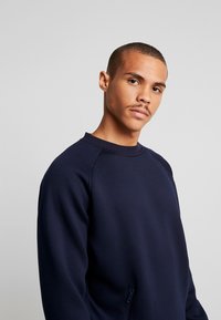 Mornarska dukserica s okruglim izrezom, raglan rukavima i bočnim detaljem sa zatvaračem. Tkanina izgleda mekano s glatkom teksturom.