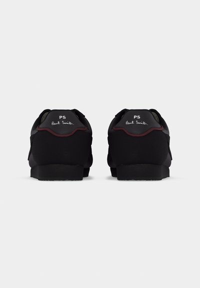 Ζευγάρι μαύρα αθλητικά παπούτσια με χαμηλές φτέρνες, με λευκή επωνυμία "PS Paul Smith" στις  πίσω ετικέτες της φτέρνας πάνω σε απλό γκρι φόντο.