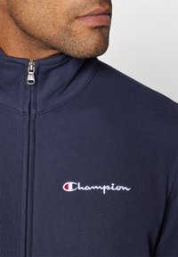 Námornícky zipsový kabát s vysokým golierom, rebrovanou textúrou a bielou vyšívanou logom "Champion" na ľavej hrudi, s striebornými doplnkami.