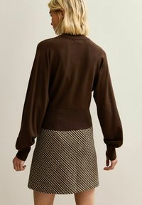 Donna con capelli biondi corti che indossa un maglione lungo marrone e una gonna a quadretti marrone e beige, rivolta verso un muro semplice.