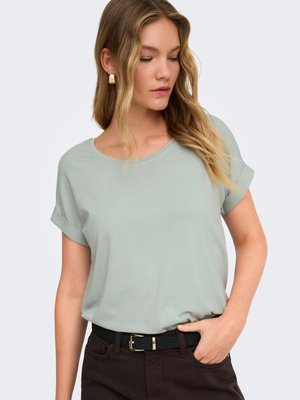 Donna con capelli biondi ondulati che indossa orecchini a cerchio dorati, una maglietta verde chiaro con maniche arrotolate, pantaloni scuri e una cintura nera con fibbia dorata.