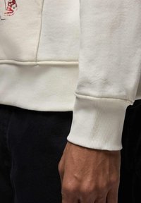 Valkoinen fleeceneule resorein hihoissa, jossa on hienovarainen painettu logo hihassa. Sileä pinta ja rento istuvuus.