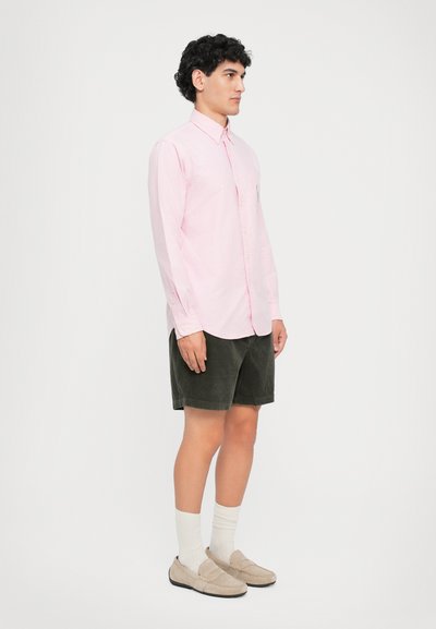 Chemise boutonnée rose clair, shorts en velours côtelé vert foncé, chaussettes blanches et chaussures à enfiler beiges. Le mannequin se tient devant un fond neutre.