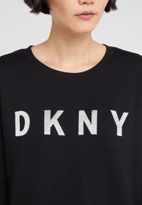 Person wearing a black sweater with large white embroidered "DKNY" letters across the chest, showing neck and lower face.