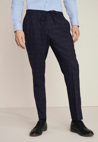 Navy geruite broek met een elastische tailleband en trekkoord, taps toelopende pasvorm en een gladde textuur. Gepaard met zwarte leren schoenen.