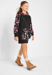 Abito felpa nero a fiori con maniche lunghe e tasca frontale. Indossato con stivali beige con lacci, calzini crema risvoltati e una piccola borsa nera.