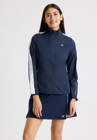 Giacca leggera blu navy con pannelli laterali bianchi, colletto alto e chiusura con zip. Presenta logo sul petto e testo sulle maniche.