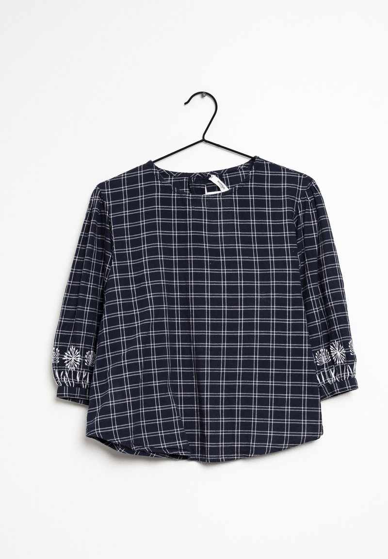 Blouse à carreaux bleu marine avec manches trois-quarts, présentant un motif de grille blanche et des accents brodés au niveau des poignets. Tissu léger.