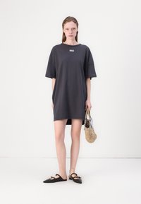 Grå oversized t-skjortekjole med korte ermer, med en liten logo. Brukt med svarte spisse leiligheter og en vevd håndveske.