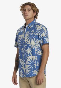 Camisa de manga corta con botonadura, que presenta un fondo azul con grandes patrones de hojas blancas. La tela tiene una textura suave y cuenta con un único bolsillo en el pecho.
