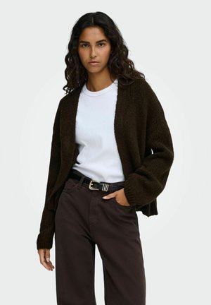 Femme aux longs cheveux bouclés portant un cardigan marron foncé, un t-shirt blanc, un pantalon marron et une ceinture noire avec une boucle dorée sur fond blanc.