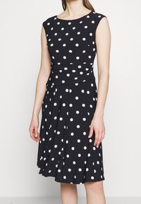 Kvinna som bär en ärmlös svart klänning med vita prickar, åtsittande midja och knälång plisserad kjol.