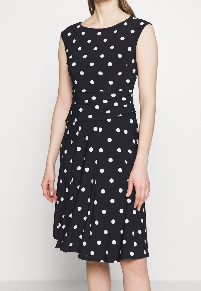 Kvinna som bär en ärmlös svart klänning med vita prickar, åtsittande midja och knälång plisserad kjol.