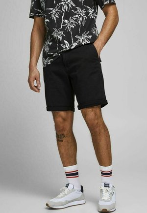 Schwarze Shorts aus Baumwollmischung, kniefrei mit umgeschlagenen Säumen; schwarz-weißes Hemd mit Palmenmotiv; graue und weiße Sneaker mit gestreiften Socken.