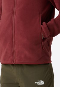 Giacca in pile rossa con zip frontale e tasca laterale. Il tessuto ha una consistenza morbida, abbinato a pantaloni verde oliva con logo.