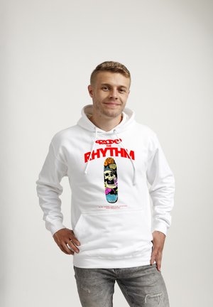 Jonge man met witte hoodie met kleurrijke skateboardafbeelding en rode tekst "Ride the Rhythm", staand met één hand op de heup.