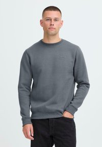 Man met kort haar in een grijze sweater met ronde hals met het "blend" logo, zwarte broek, staand met één hand in de zak tegen een witte achtergrond.