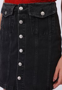 Jupe en denim noir avec fermeture à boutons sur le devant, deux poches avant et détails de couture verticale. Texture lisse avec des boutons métalliques.