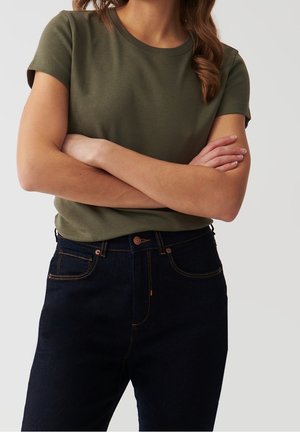 Oliwkowo-zielona bawełniana koszulka z krótkim rękawem, zestawiona z ciemnoniebieskimi dżinsami z pomarańczowym przeszyciem i zapinanym na guziki pasem.