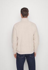 Chaqueta de forro polar de color beige claro con un cuello alto, puños elásticos y dobladillo. La textura es suave y esponjosa, ofreciendo un aspecto acogedor.
