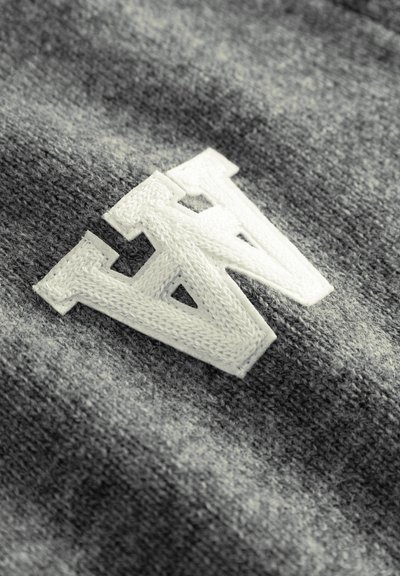 Sivá tkanina s textúrovaným povrchom, na ktorom dominuje biele vyšívané písmeno "N". Detailný pohľad zdôrazňuje detaily stehu.