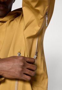 Żółta wodoodporna kurtka ma dwa boczne zamki, gładką fakturę i luźny krój. Zbliżenie na rękę regulującą zamek.