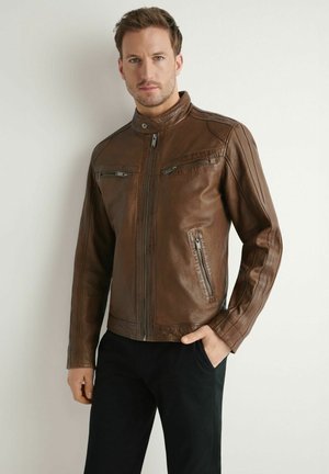 Mężczyzna w brązowej skórzanej kurtce z zamykanymi kieszeniami, stoi z jedną ręką w kieszeni na jednolitym jasnym tle.