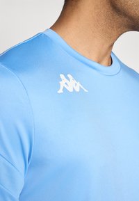 Šviesiai mėlyna sportinė marškinė, pagamintos iš lygaus audinio, su apvalia iškirpte ir baltu logotipu kairėje krūtinės pusėje.