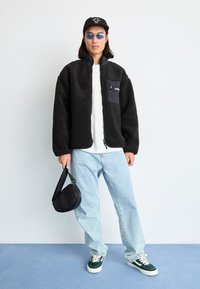 Flauschige schwarze Fleecejacke mit Reißverschluss und Brusttasche, kombiniert mit hellblauen Jeans und grünen Schuhen. Accessoires sind eine schwarze Tasche und Sonnenbrille.